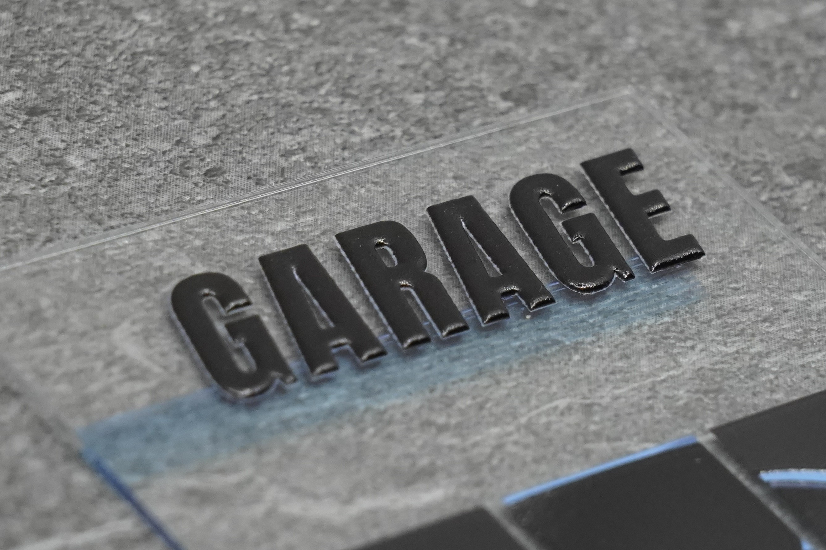 Makro-Aufnahme der erhaben gedruckten Buchstaben «GARAGE» auf transparentem Acryl — Buchstabenhöhe wenige Millimeter, sichtbare 3D-Prägung mit glänzender Oberfläche
